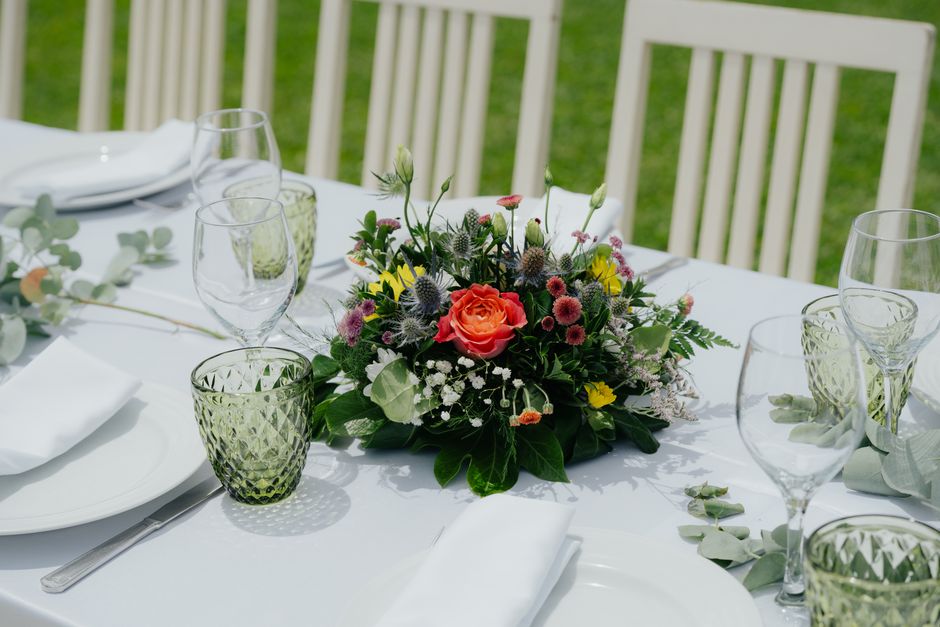 A floral centerpiece featuring a vibrant bouquet of fresh flowers, including a prominent pink-orange rose, surrounded by small pink, purple, and yellow flowers, along with green foliage. The arrangeme