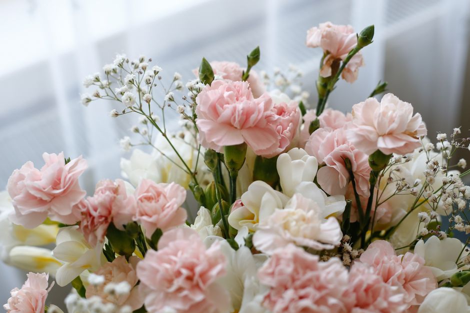 A close-up of a floral arrangement featuring soft pink carnations and white roses, accented with delicate sprigs of baby's breath. The flowers are arranged in a clear glass vase, highlighting their fr