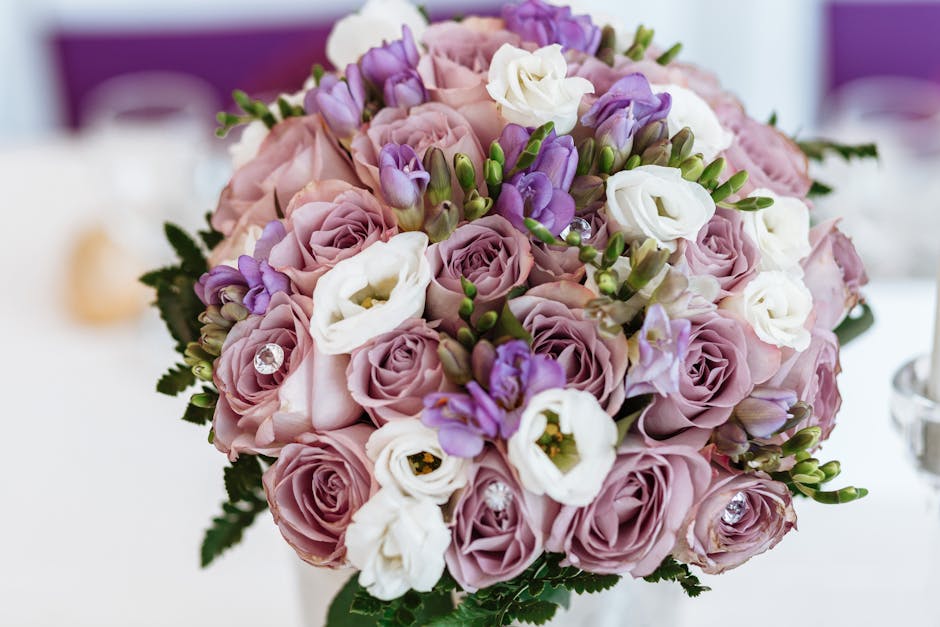 A close-up of a floral bouquet featuring pale pink roses, white lisianthus, and purple freesias, densely arranged with fresh, vibrant petals and green foliage. The flowers are artistically clustered a