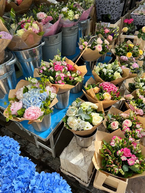 A display of freshly prepared floral bouquets arranged in metal buckets and paper wraps, featuring a variety of flowers including pink roses, white freesias, blue hydrangeas, and carnations. The bouqu