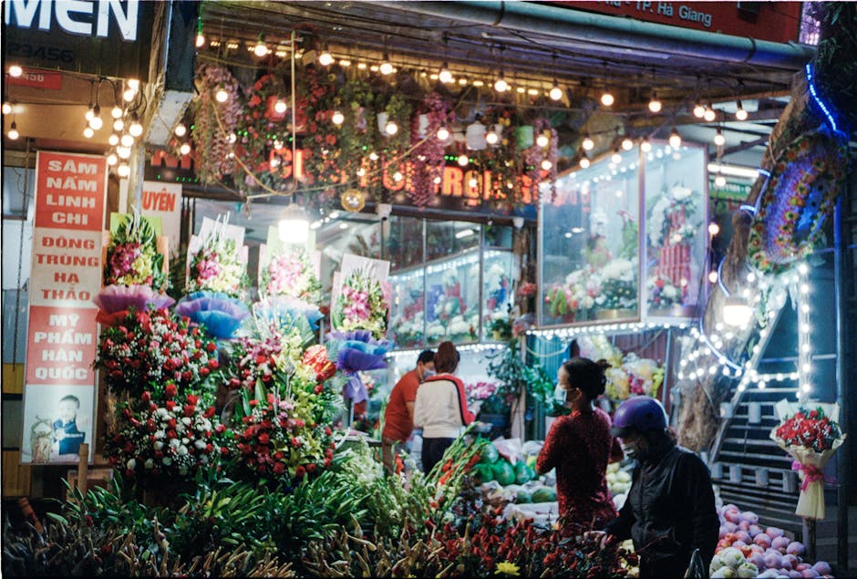 A vibrant flower market scene illuminated by colorful string lights, featuring a variety of fresh blooms arranged in bouquets and floral displays. In the foreground, there are lush red and white roses