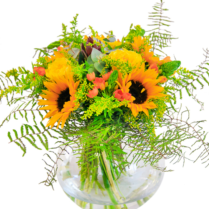 The first impression of this radiant bouquet is pure happiness-a vivid spectrum of yellow, orange, and green hand-delivered with the artistry Forest Gate is known for. At its heart are several exuberant sunflowers, their golden crowns circling dark, velvet-like centers, conjuring up memories of sunny allotment gardens and strolls through Wanstead Park. Against their bold presence, buttery yellow roses unfold in soft, choreographed spirals, their delicate scent rising with every petal. Glossy red Hypericum berries cluster playfully among the blooms, providing flashes of color reminiscent of ripening fruit at local Forest Gate markets. Generous wisps of fern and solidago, bright with tiny yellow blossoms, cascade and curl at the edges, evoking the gentle wildness of East London's green spaces. Subtle hints of blue-green eucalyptus sneak through the foliage, grounding the design with their silvery hue and faintly refreshing aroma. The bouquet rests in a clear, globe-shaped vase that reveals water-beaded stems, adding a sense of freshness and architectural grace. The arrangement is alive with texture-the downy surface of sunflowers, tightly wound rose petals, glossy berries, and the crisp snap of new leaves-each part harmonizing to form a whole that is both uplifting and soothing. Designed for joyous milestones or just-because days, this bouquet reflects the heart and seasonality of Forest Gate Flowers, delivering a splash of light and celebration to any home, from Upton Lane to Maryland Point.