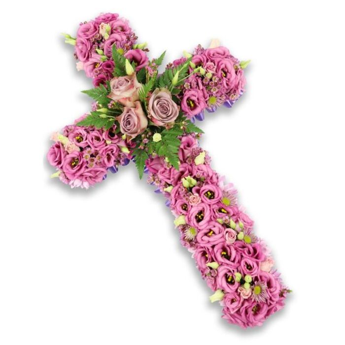 Against a bright white background, a meticulously crafted floral cross seems to float, its delicate shadow emphasizing every curve of the design. The structure is completely covered in flowers, so closely arranged that the surface appears almost quilted with petals. At the very centre where the arms of the cross meet, three large roses in muted dusty rose and light mauve tones rest together like a quiet cluster of memories. Their petals are generously layered and fully open, revealing soft spirals that draw the gaze inward. Around these central roses, feathery green fern fronds and smaller glossy leaves provide a fresh contrast, creating pockets of depth that make the arrangement feel alive. Stretching out from this central point, the arms of the cross are filled with lisianthus blossoms in various shades of pink, lilac, and delicate purple. Some of these trumpet-shaped blooms display deeper, darker centres, adding a sophisticated, almost painterly effect. Tiny accent flowers in pink and violet, along with pale cream and white blossoms with subtle yellow centres, are scattered thoughtfully throughout, like whispered notes of light. Here and there, slivers of deeper purple ribbon or fabric peek from beneath the densely packed flowers at the edges, hinting at the skilled construction behind the beauty. Designed in the spirit of a Forest Gate farewell, this floral cross would feel at home in a quiet chapel off Woodgrange Road or during a graveside service, offering a serene, faith-filled tribute. It carries a message of respect, peace, and remembrance for a loved one whose life is being gently honoured in flowers.